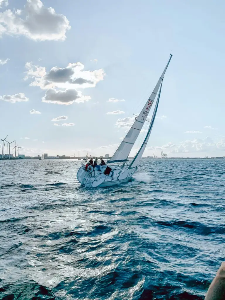 A sailboat glides through Copenhagen's waters against a backdrop of wind turbines under a bright sky.